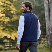 Man in a Tom Beckbe Warren Fleece Vest over a plaid shirt, walking outdoors with trees and a fence behind him.