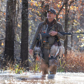 A man in a Tom Beckbe DU Waterfowl Strap Vest stands in shallow water with a rifle and ducks, surrounded by autumn trees.
