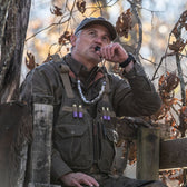 Man in a Tom Beckbe DU Waterfowl Strap Vest blows a duck call on a wooden platform amid dry autumn leaves.
