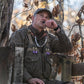Man in a Tom Beckbe DU Waterfowl Strap Vest blows a duck call on a wooden platform amid dry autumn leaves.