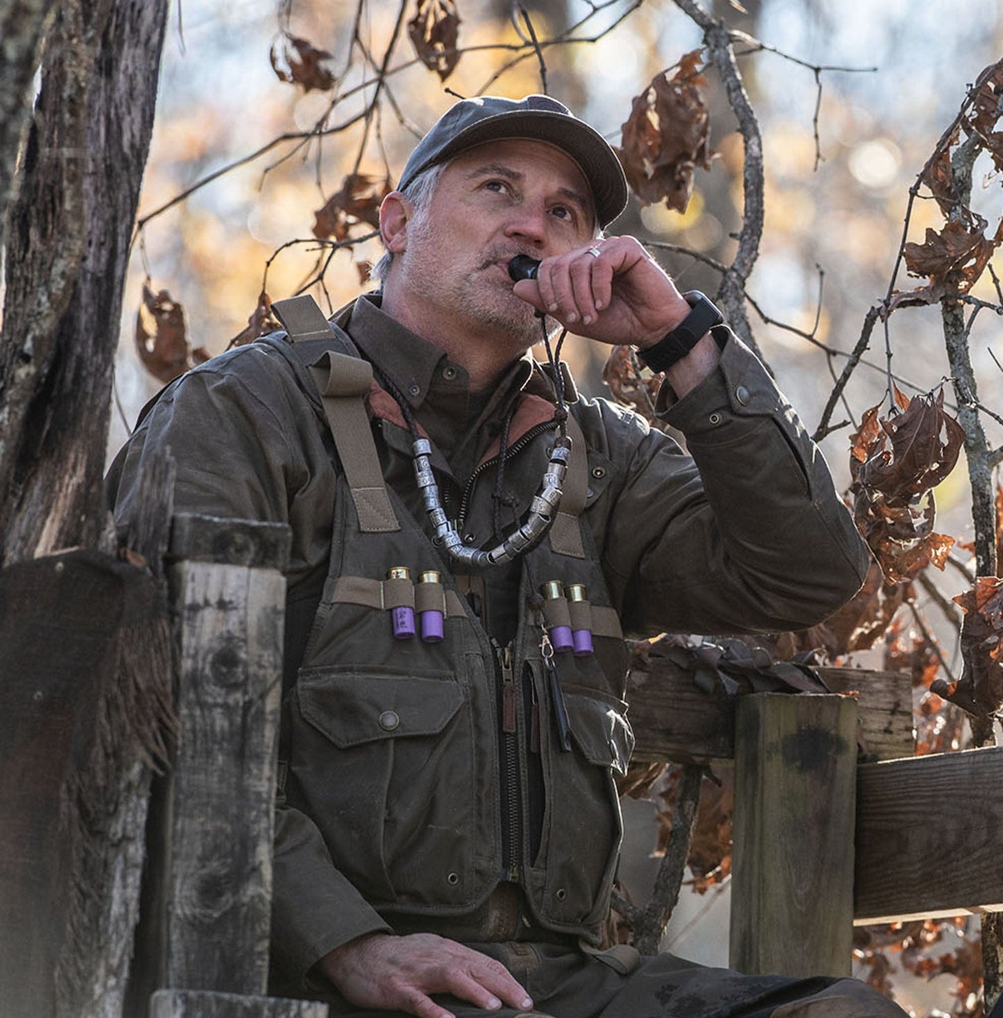 Man in a Tom Beckbe DU Waterfowl Strap Vest blows a duck call on a wooden platform amid dry autumn leaves.