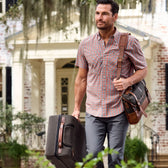 Man with a plaid shirt and Tom Beckbe Canvas Wheeled Carry-On Bag stands before a brick house with a white door.