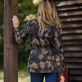 A person with long blonde hair stands outside, facing away from the camera, wearing Tom Beckbes Womens Blakeley Jacket in a camo pattern and blue jeans. One hand rests on a wooden post as the jackets weatherproof cotton shell complements the rustic wooden building and lush greenery in the background.