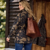 A woman with long blonde hair wears the Tom Beckbe Womens Blakeley Jacket and blue jeans. She carries a brown leather tote over her shoulder, standing outside a wooden building, gazing left. A hanging coat in the background completes her feminine silhouette.