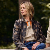 A person wearing a Tom Beckbe Womens Blakeley Jacket over a beige shirt and blue jeans sits outdoors. With shoulder-length blonde hair, they are looking to their right against a blurred leafy green backdrop that captures natures tranquility.