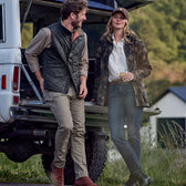 A man and woman lean against an open truck tailgate. The man wears a quilted vest and plaid shirt, while the woman sports the Tom Beckbe Womens Blakeley Jacket and a baseball cap. They appear relaxed, enjoying a drink in the countryside setting.