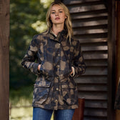 A woman stands outdoors in a Tom Beckbe Womens Blakeley Jacket with a camo pattern, paired with jeans. She leans against a wooden structure, surrounded by blurred greenery, with her hands in her pockets.