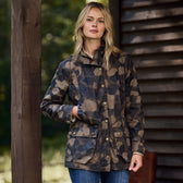 Standing outdoors by a wooden structure with a forest backdrop, a person in Tom Beckbes Womens Blakeley Jacket and jeans exudes natural flair. With hands in their jacket pockets and long blonde hair cascading down, the waxed camouflage attire perfectly balances the feminine silhouette.