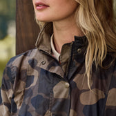 A person with long, wavy blonde hair sports the Womens Blakeley Jacket by Tom Beckbe, collar up. The weatherproof cotton shell subtly glistens with a zipper and snap buttons. Lips and chin peek through as the softly blurred background suggests greenery.