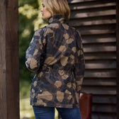 A blonde person stands outdoors, wearing a Womens Blakeley Jacket by Tom Beckbe that highlights a feminine silhouette. They face away, with a wooden building to their right and greenery in the background.