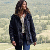 A woman in a Tom Beckbe Womens Blakeley Jacket and jeans walks outdoors, with a green landscape in the background.