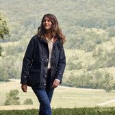A person strolls on a grassy field wearing Tom Beckbes Womens Blakeley Jacket and jeans, with hills in the scenic background.