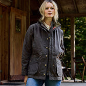 A person with blonde hair stands outside a wooden cabin, wearing a Tom Beckbe Womens Blakeley Jacket and blue jeans. The background features trees and chairs.