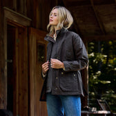On a wooden porch, a person in blue jeans models the Tom Beckbe Womens Blakeley Jacket. With shoulder-length wavy blonde hair, they look upward. A rustic wooden building framed by trees serves as the backdrop.