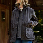 A blonde-haired individual is wearing a Tom Beckbe Womens Blakeley Jacket with a high collar and has their hands in their pockets while standing before a wooden structure and a blurred green backdrop, paired with blue jeans.