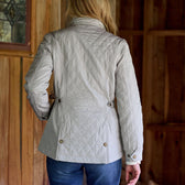 Person in Tom Beckbe Womens Camden Quilted Jacket and jeans, standing with back turned in a wooden setting.