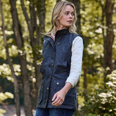 A woman in a Tom Beckbe Womens Fairmont Vest and white shirt stands looking sideways outdoors in a forest.