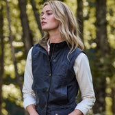 In a forest, a person in the Tom Beckbe Womens Fairmont Vest over a light shirt gazes sideways with trees blurred behind.