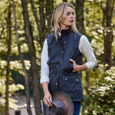 A woman stands in a wooded area, wearing a white shirt and Tom Beckbes Womens Fairmont Vest, holding a hat in her left hand.