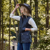 A woman wearing a Tom Beckbe weatherproof hat and Womens Fairmont Vest stands in a forest, gazing to the side.