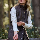 Woman in Tom Beckbe Womens Fairmont Vest over a white long-sleeve shirt adjusts sleeve, with a forested backdrop.
