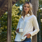 Wearing a Tom Beckbe Womens Oxford Shirt, she holds a mug outdoors near a wooden structure with trees behind her.
