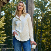 A woman in a Tom Beckbe Womens Oxford Shirt and jeans holds a mug by a wooden post with trees behind her.