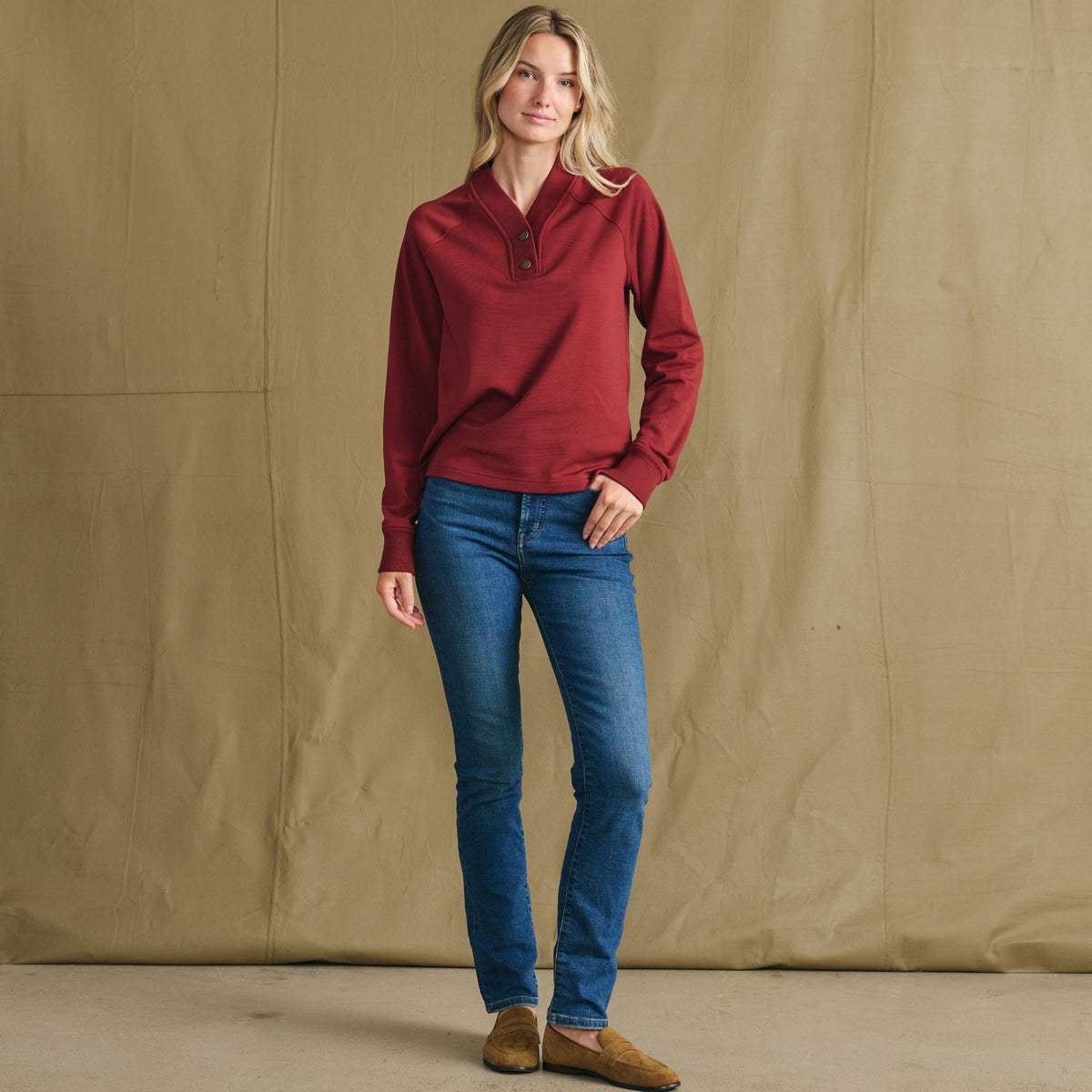 Woman poses before tan backdrop in Tom Beckbe Women’s Shelby Fleece Pullover, blue jeans, and brown shoes.