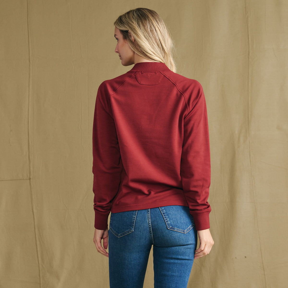 Woman wears Tom Beckbe Women’s Shelby Fleece Pullover in red and blue jeans, facing away from a beige fabric backdrop.
