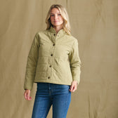 She wears a Tom Beckbe Women’s Tyler Quilted Jacket and blue jeans, standing before a plain beige background.