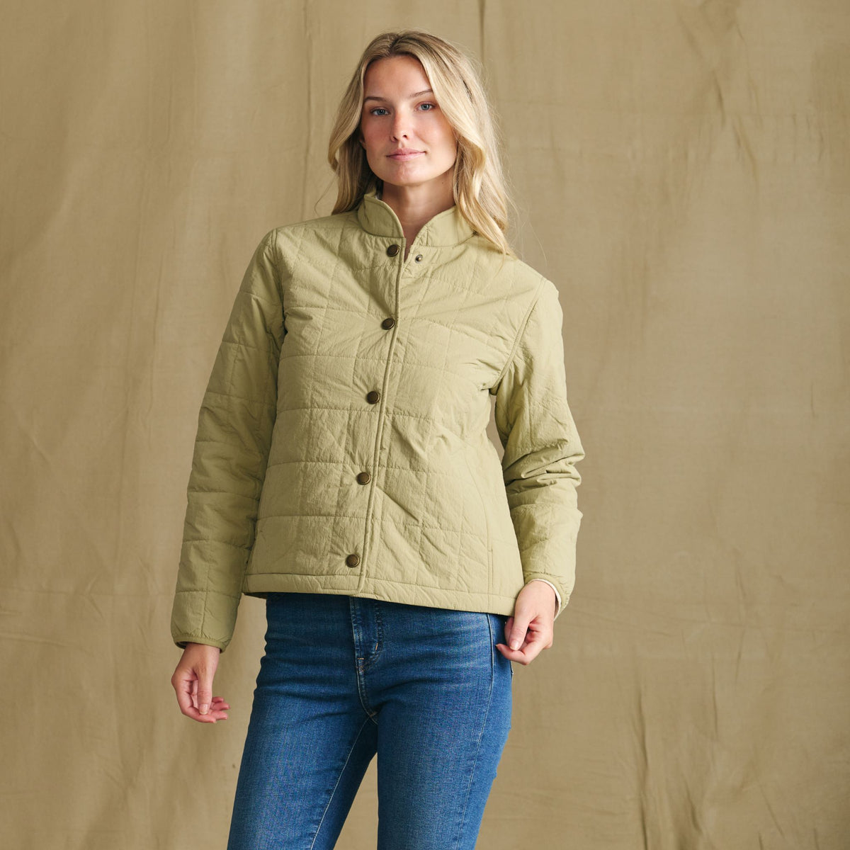 She wears a Tom Beckbe Women’s Tyler Quilted Jacket and blue jeans, standing before a plain beige background.