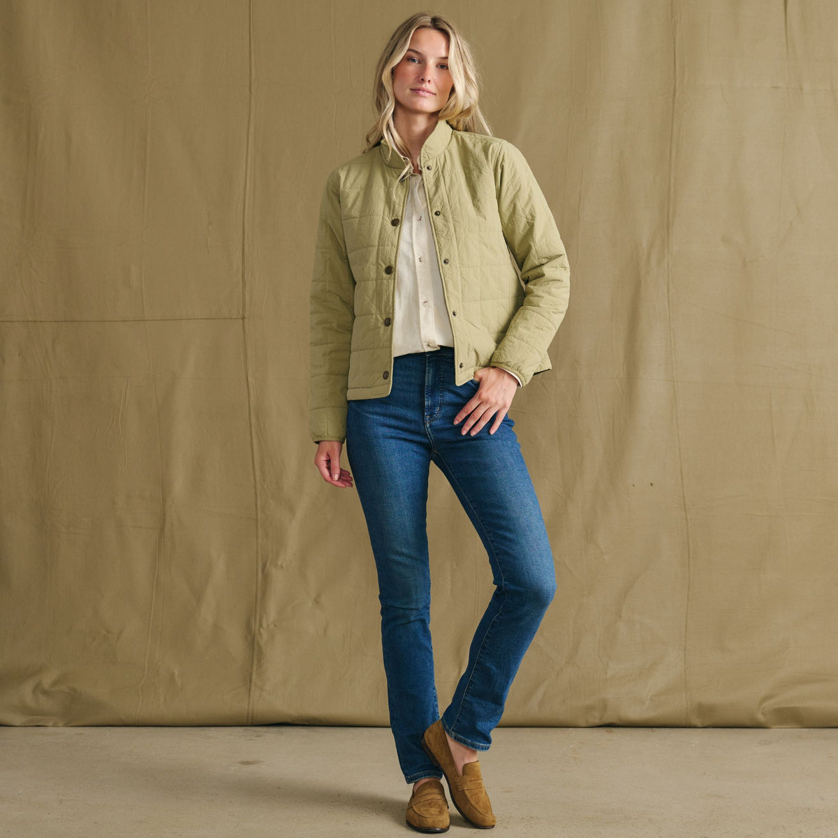 Indoors, a woman wears Tom Beckbe’s Women’s Tyler Quilted Jacket, blue jeans, tan shoes, and a cream shirt.