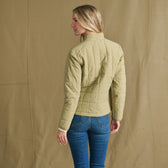 A woman faces away, modeling the Tom Beckbe Women’s Tyler Quilted Jacket with blue jeans against a tan backdrop.