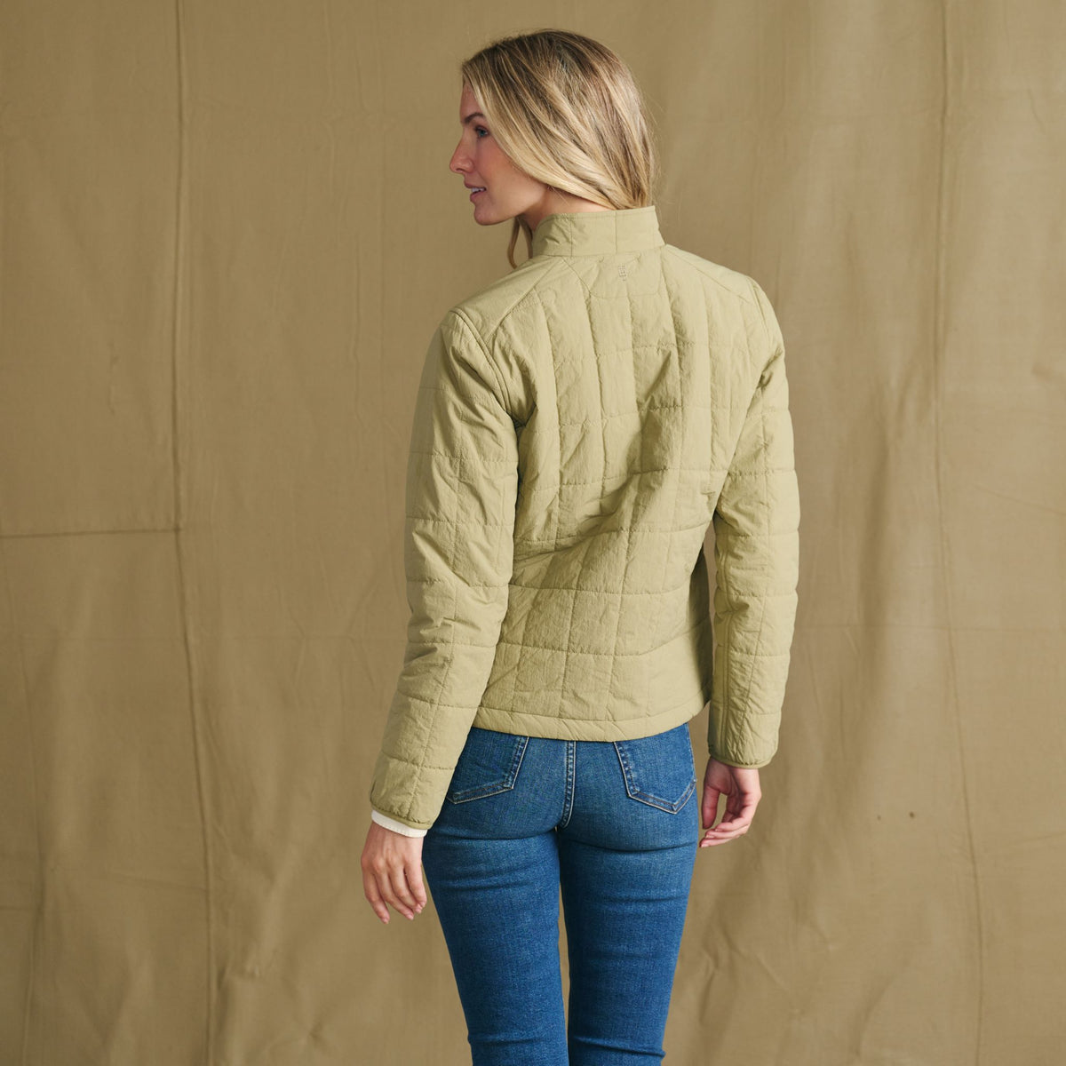 A woman faces away, modeling the Tom Beckbe Women’s Tyler Quilted Jacket with blue jeans against a tan backdrop.
