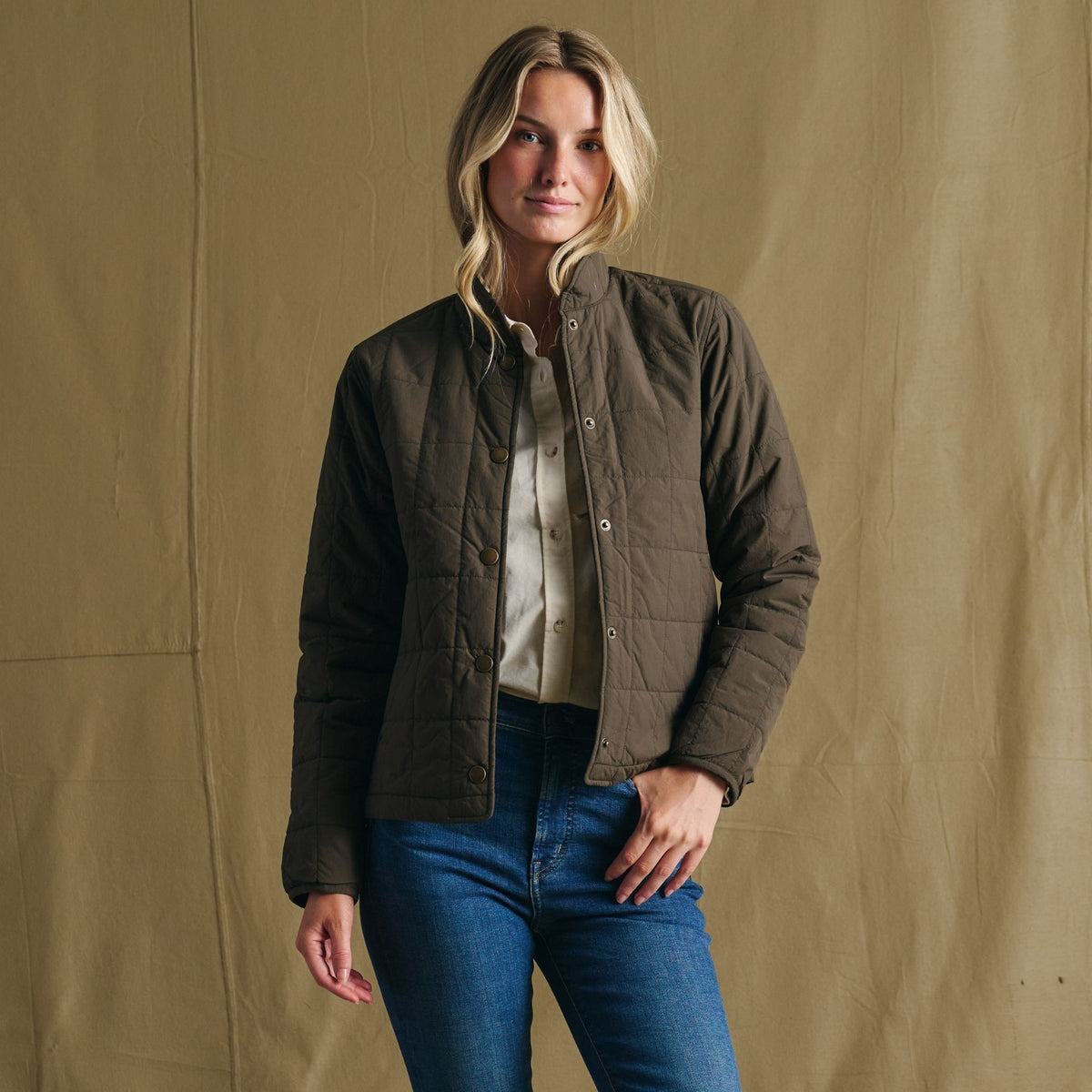 Woman in Tom Beckbe Women’s Tyler Quilted Jacket, light shirt, and jeans stands before a beige fabric backdrop.