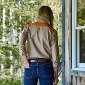 A person in a tan Womens Uplander Shirt by Tom Beckbe and blue jeans stands near a wooden structure.