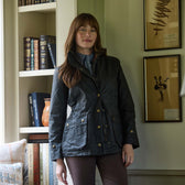 A woman in a dark jacket stands indoors near a bookshelf and framed botanical prints on the wall.