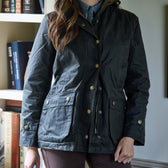 Person wearing a dark utility jacket over a blue shirt, standing indoors next to a bookshelf.