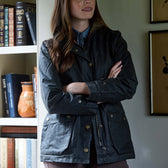 Woman in a dark jacket stands with arms crossed near a bookshelf and framed art, facing slightly away from the camera.