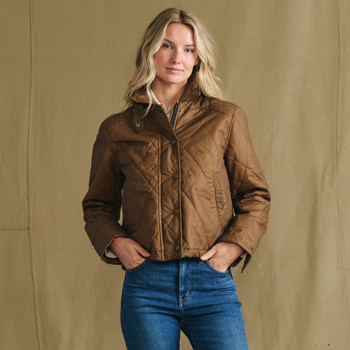 Woman wearing a brown quilted jacket and blue jeans stands in front of a plain tan background with hands in pockets.