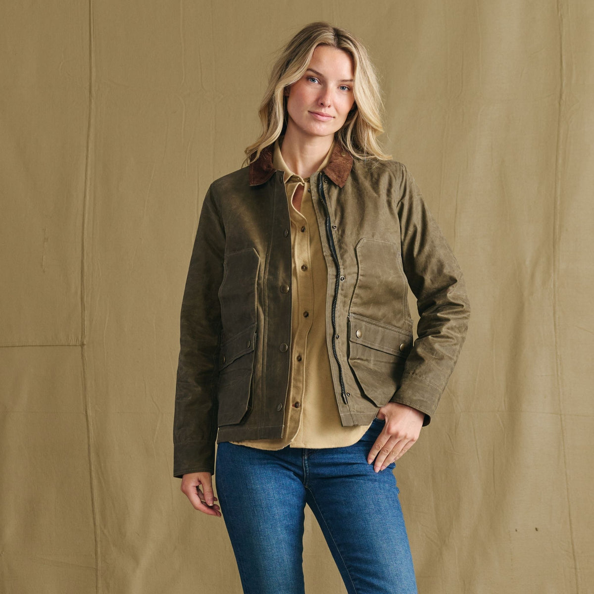 Woman wears Tom Beckbe Women’s Steward Jacket with blue jeans, standing against a tan backdrop, facing the camera.