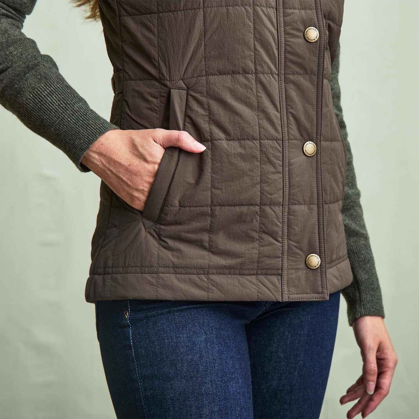 Woman wearing a Tom Beckbe Womens Tyler Quilted Vest with a green sweater and blue jeans, hand in vest pocket.