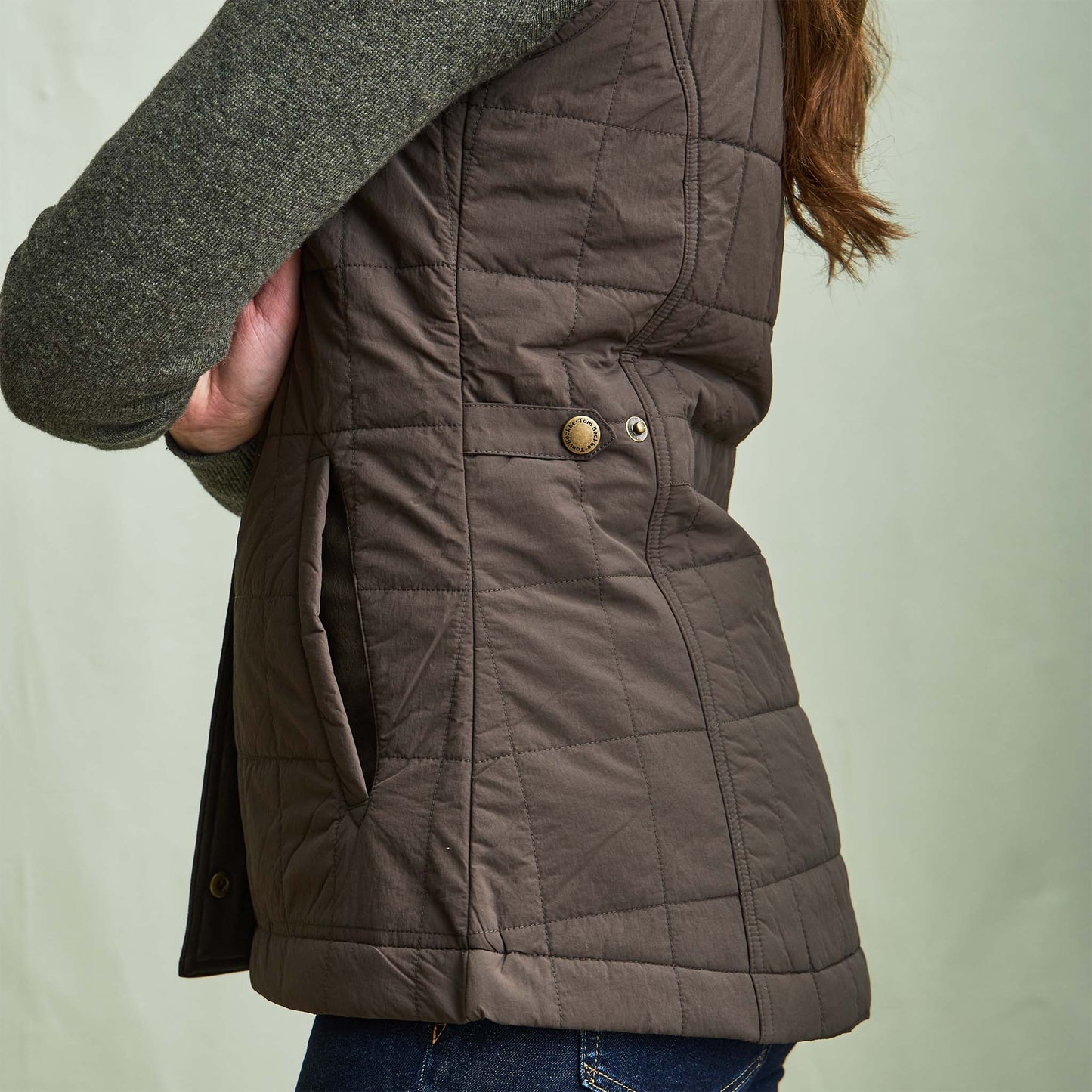 Woman wearing a Tom Beckbe Womens Tyler Quilted Vest over a grey long-sleeve shirt and blue jeans, shown from the side.