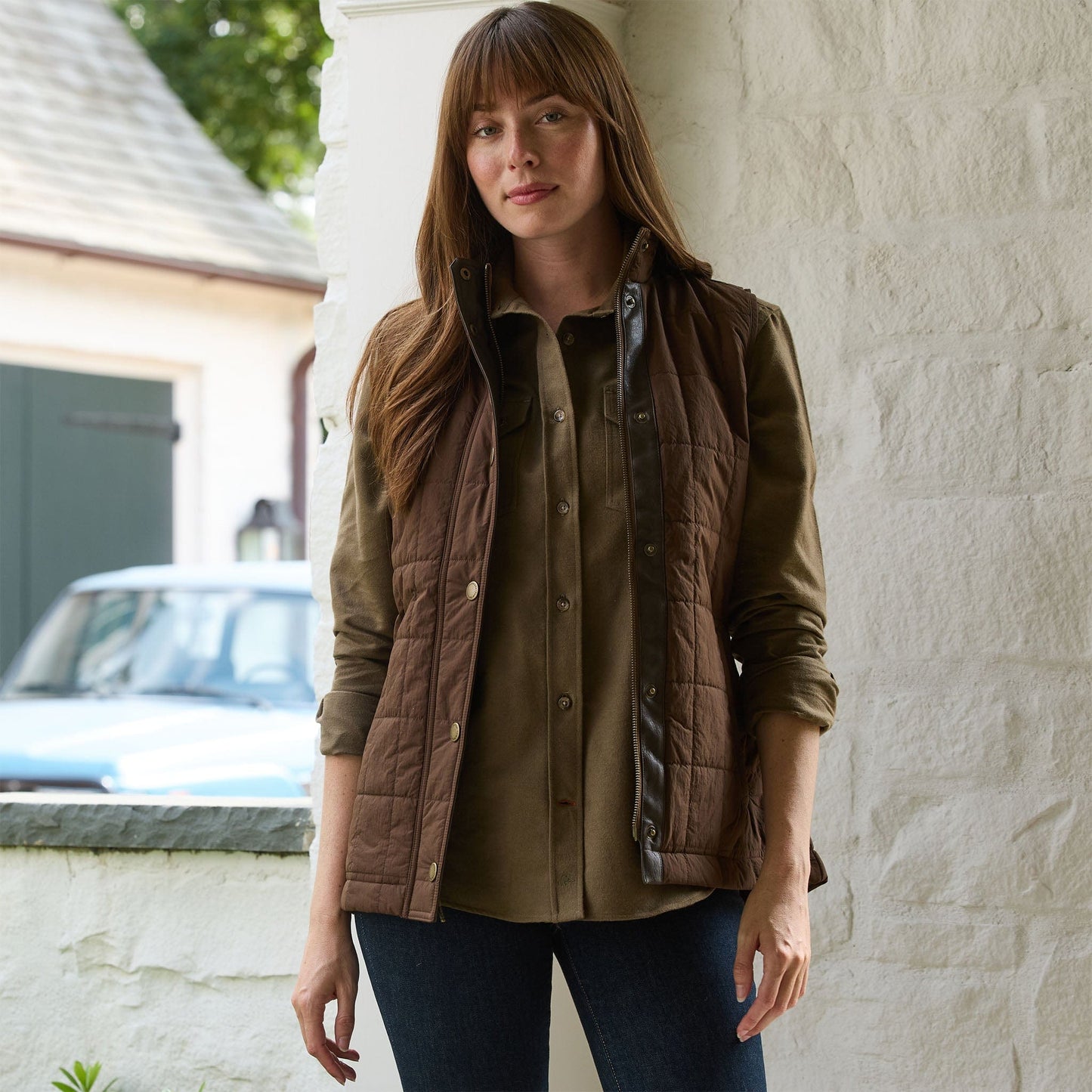 Woman outside in a Tom Beckbe Womens Tyler Quilted Vest over a buttoned shirt and dark jeans by a white wall.