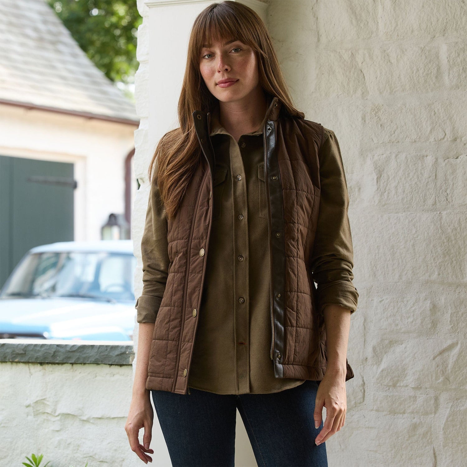 Woman outside in a Tom Beckbe Womens Tyler Quilted Vest over a buttoned shirt and dark jeans by a white wall.