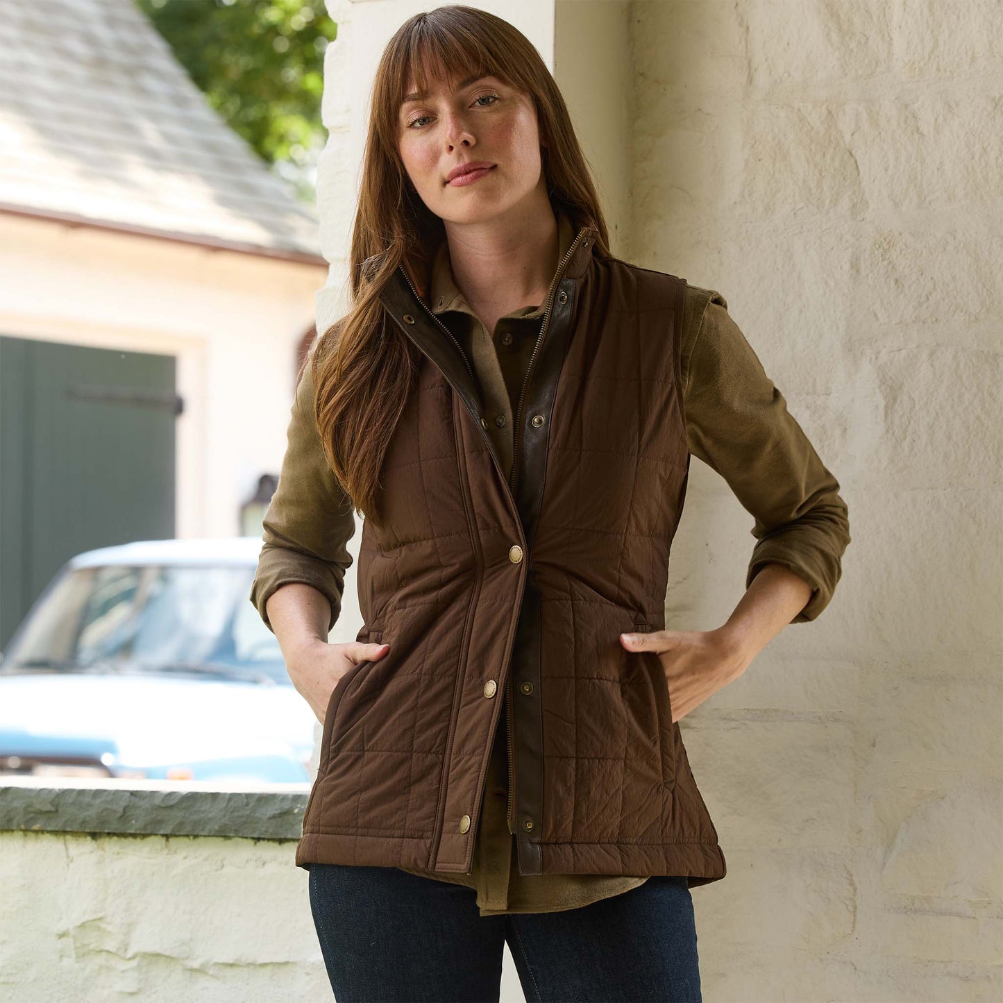 A woman wears the Tom Beckbe Womens Tyler Quilted Vest over a green shirt, standing by a white wall with hands in pockets.