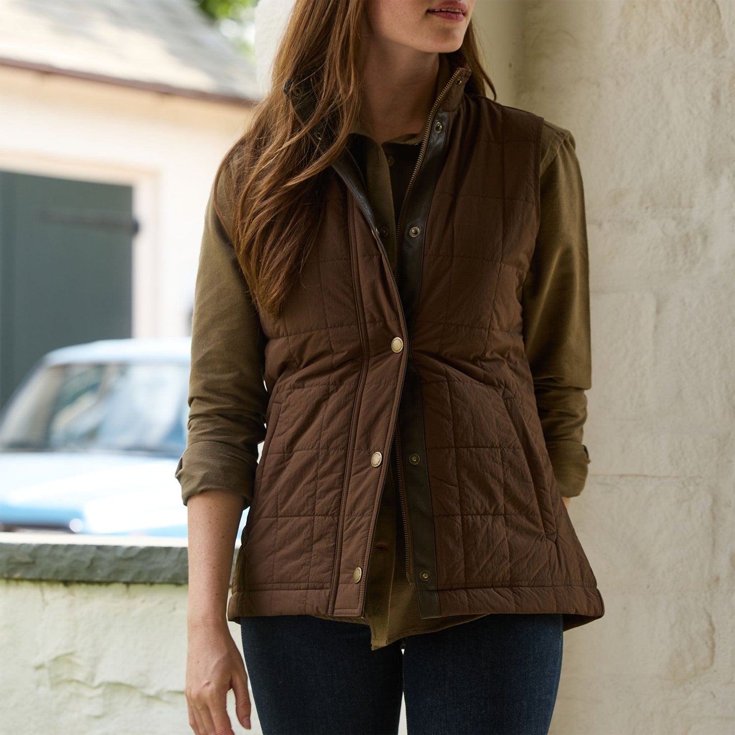 Woman wearing the Tom Beckbe Womens Tyler Quilted Vest over a green shirt and jeans, standing by a white wall outdoors.