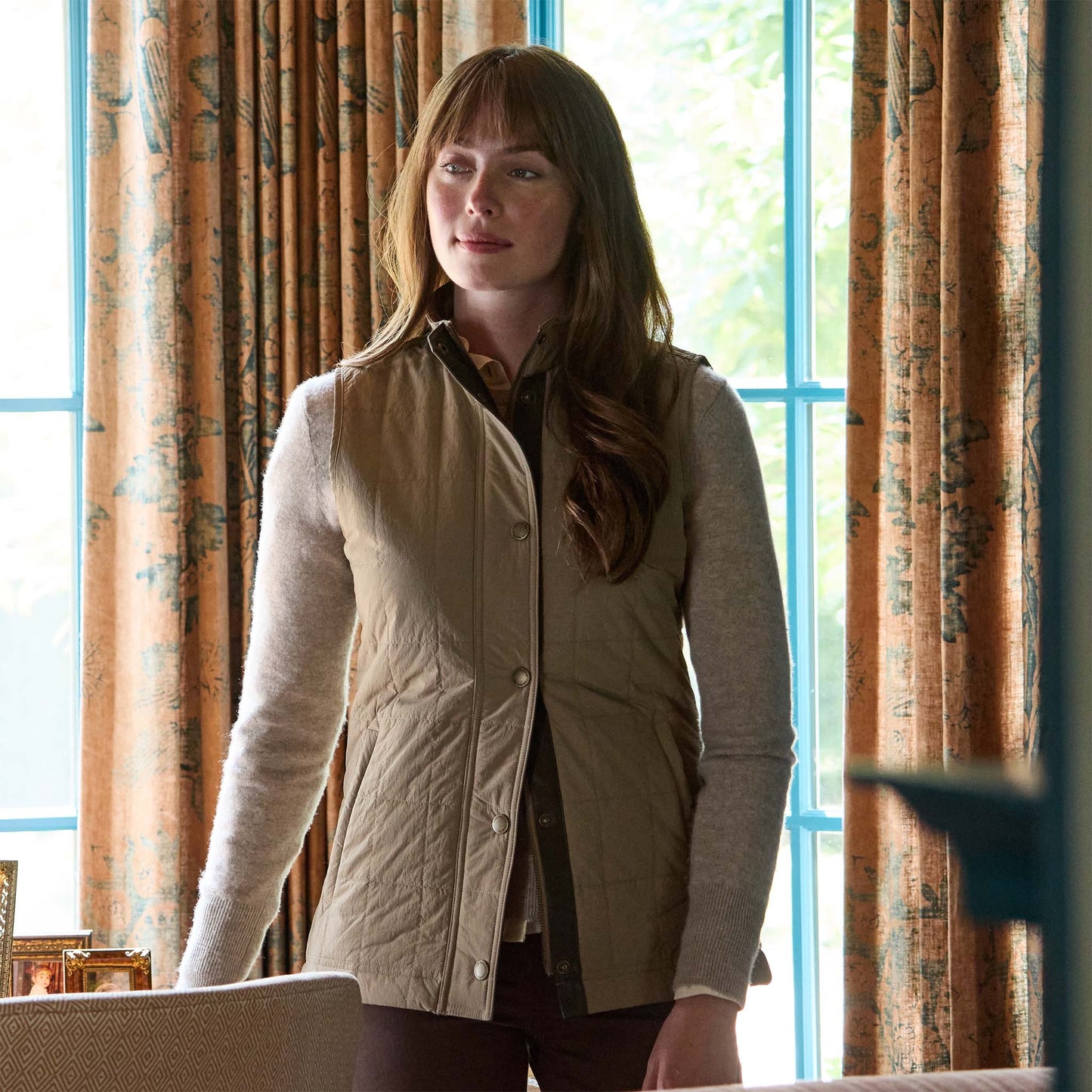A woman with long brown hair wears the Tom Beckbe Womens Tyler Quilted Vest indoors near patterned curtains and a window.