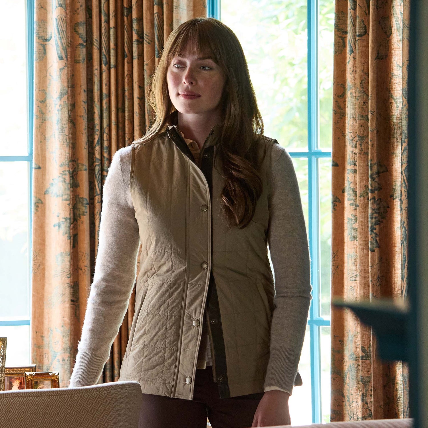 A woman with long brown hair wears the Tom Beckbe Womens Tyler Quilted Vest indoors near patterned curtains and a window.