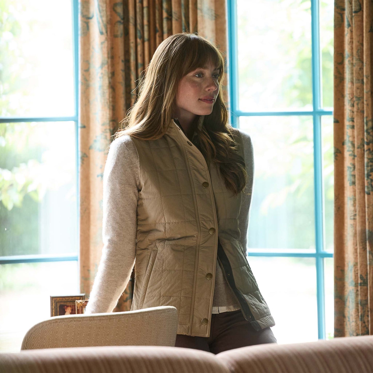 A woman with long hair wears the Tom Beckbe Womens Tyler Quilted Vest indoors by a sunlit window with patterned curtains.